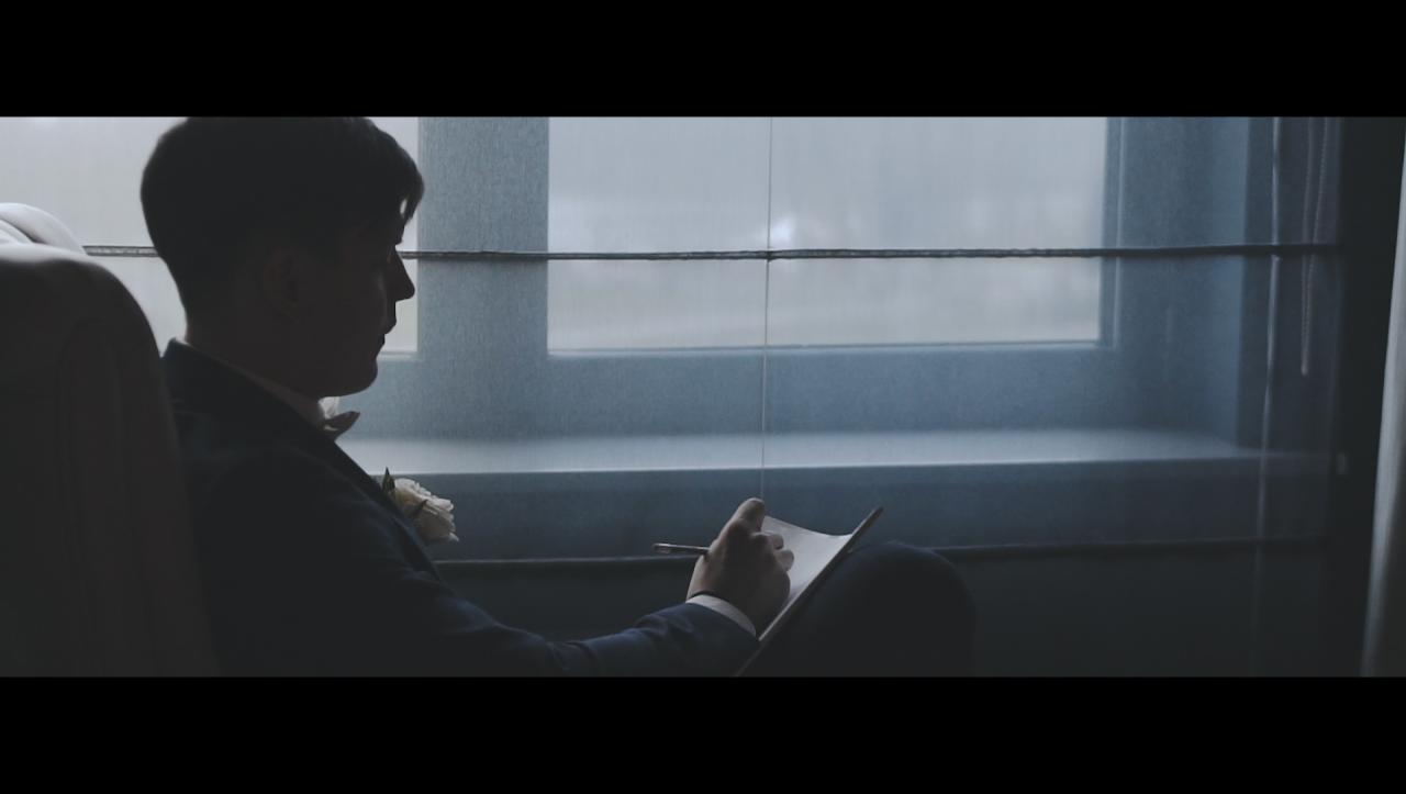 Mężczyzna w ciemnym garniturze z butonierką i muszką, siedzi przy oknie i pisze coś na kartce, prawdopodobnie przemówienie weselne.