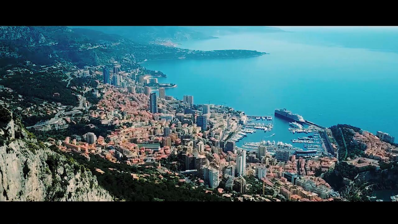 Panoramiczny widok z góry na Monako: gęsta zabudowa miejska z wieżowcami, port jachtowy z zacumowanym dużym wycieczkowcem, błękitne morze i zalesione góry w tle.