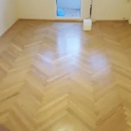 Parkiet ułożony w jodełkę, po cyklinowaniu, z widocznym pojemnikiem z lakierem i wałkiem do lakierowania, w jasnym pomieszczeniu z oknem.