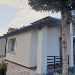 Kamox - Jasny, świeżo otynkowany budynek z elewacją natryskową, widoczna drabina oparta o ścianę, kwiaty w donicach na pierwszym planie, fragment drzewa po prawej stronie.