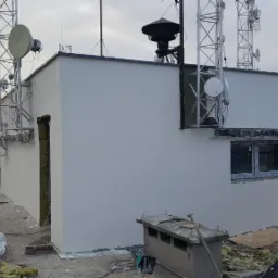 Świeżo pomalowana elewacja budynku technicznego na dachu w Stargardzie, z widocznymi antenami telekomunikacyjnymi i elementami wykończeniowymi okna, w trakcie prac porządkowych.