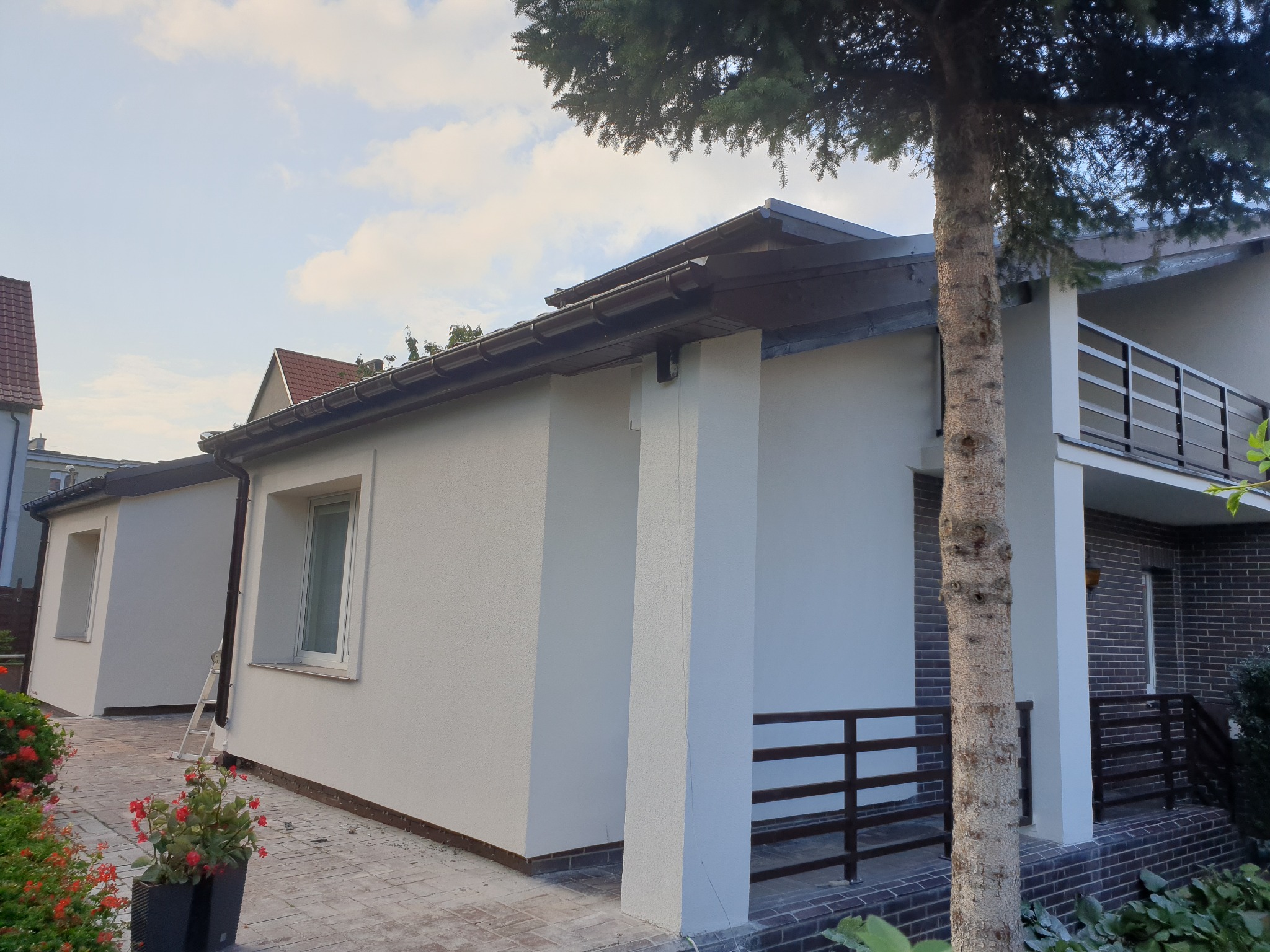 Jasny, świeżo otynkowany budynek z elewacją natryskową, widoczna drabina oparta o ścianę, kwiaty w donicach na pierwszym planie, fragment drzewa po prawej stronie.