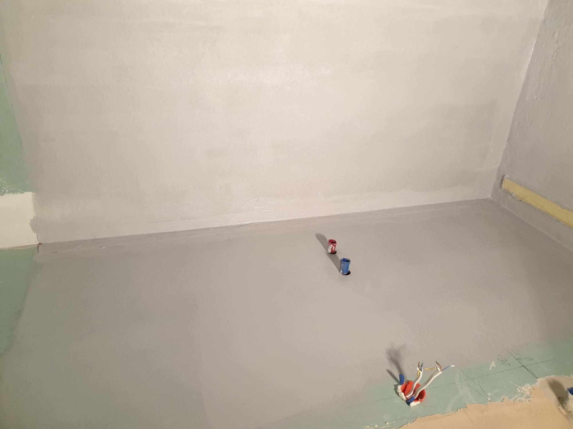 Szare hydroizolacja w rogu łazienki, widoczne wystające rury i kable elektryczne, przygotowanie do prac wykończeniowych.