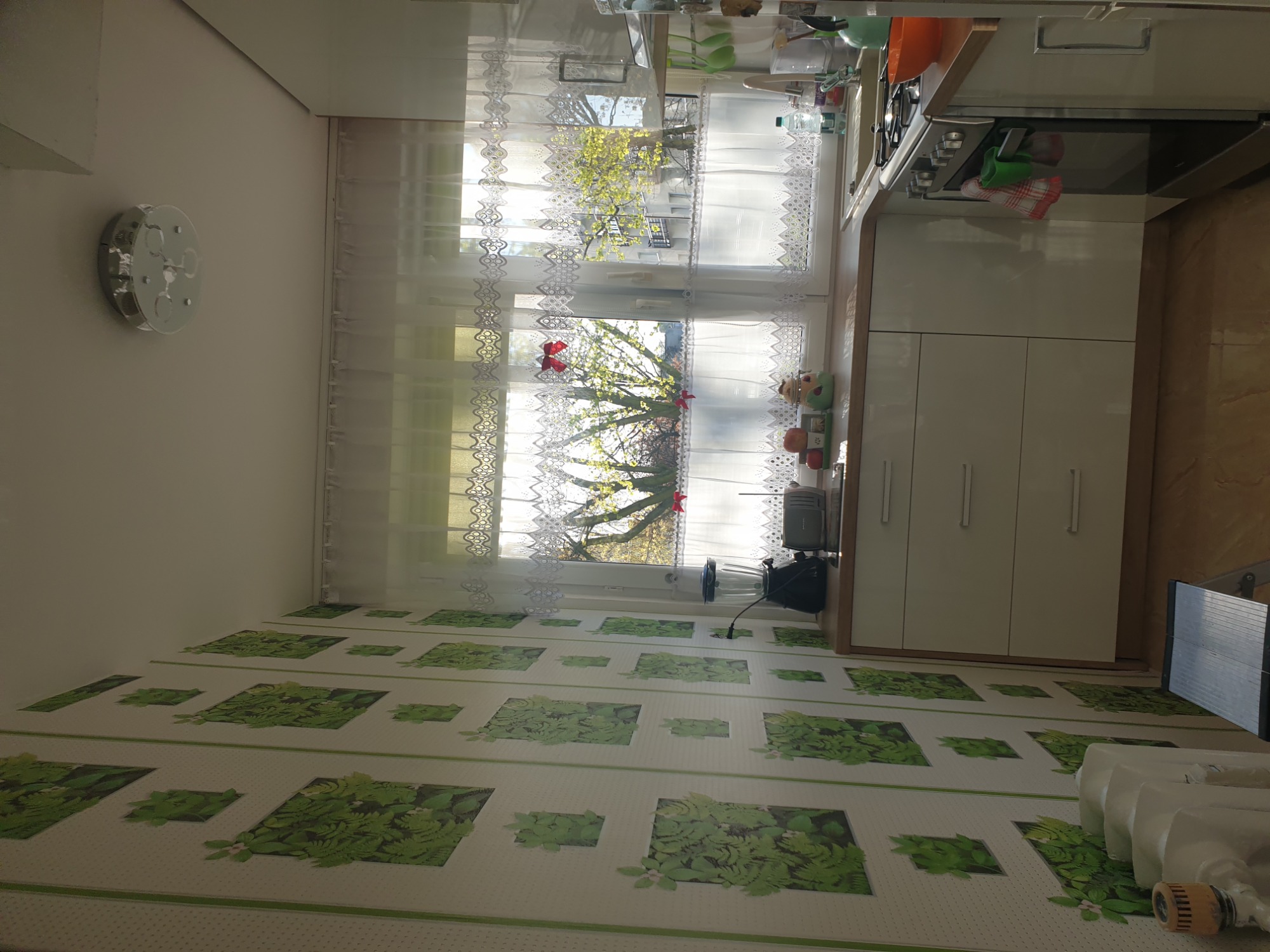 Jasna kuchnia z widokiem na drzewo przez okno z koronkowymi firankami, na ścianie wzorzysta tapeta z motywem liści, widoczny fragment białych szafek kuchennych i sprzęt AGD.