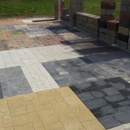 ARCO - MATERIAŁY BUDOWLANE Arkadiusz Zyśk - Ekspozycja różnych wzorów kostki brukowej ułożonych obok siebie, prezentujących różnorodność kolorów i faktur, z fragmentem murku w tle.