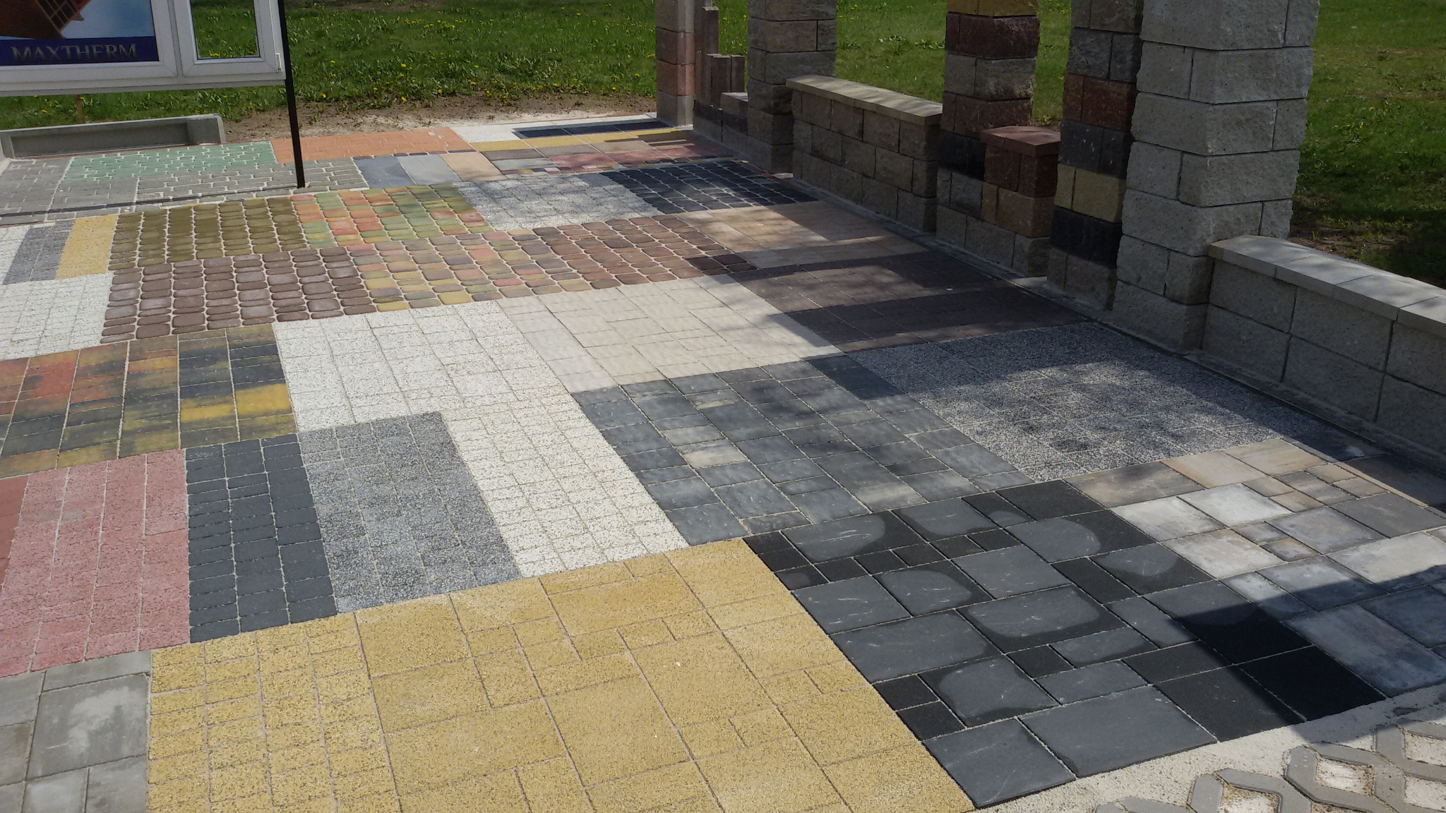 Ekspozycja różnych wzorów kostki brukowej ułożonych obok siebie, prezentujących różnorodność kolorów i faktur, z fragmentem murku w tle.