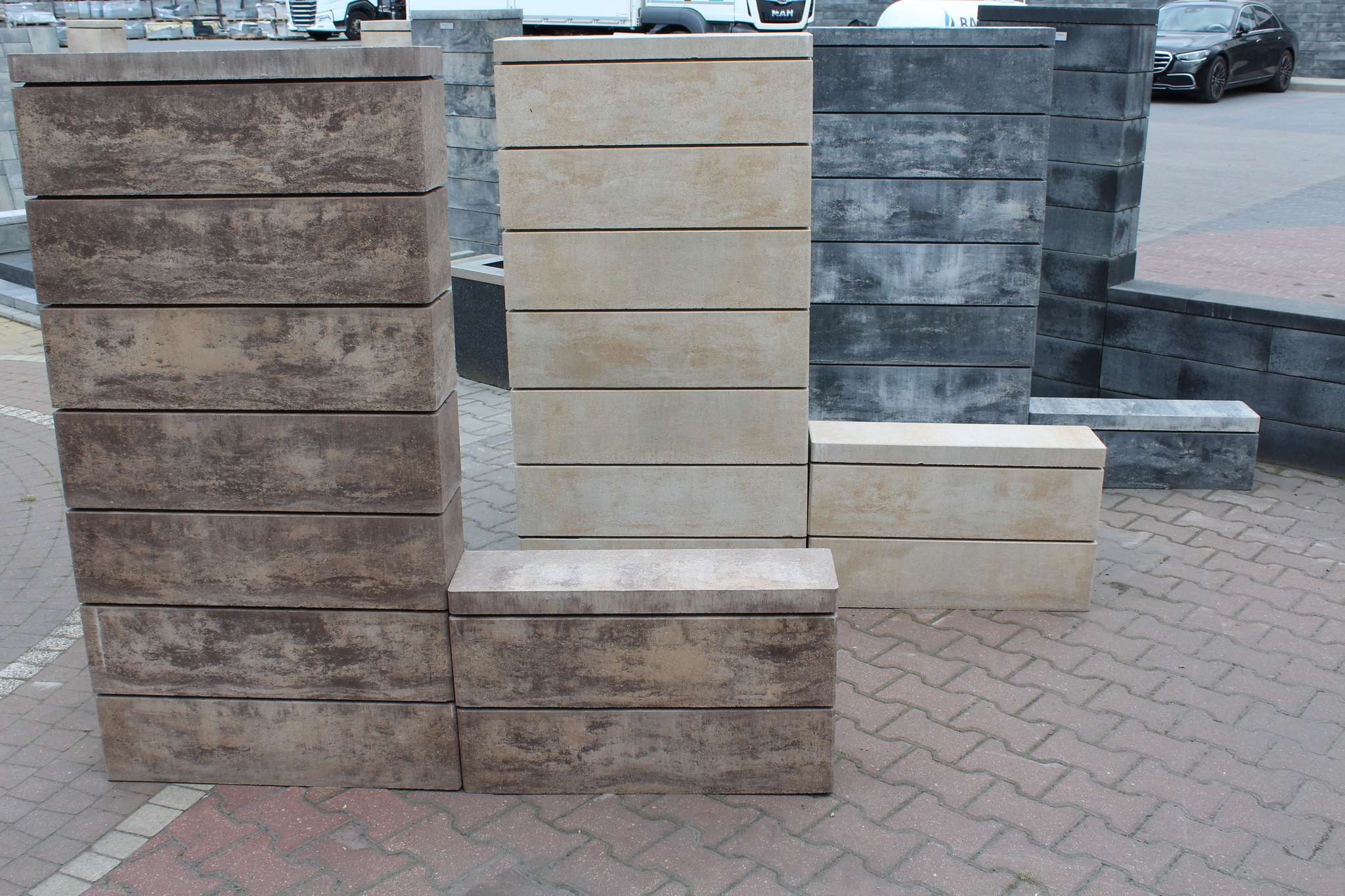 Ułożone kostki betonowe w trzech różnych kolorach i fakturach: brązowe, beżowe i grafitowe, na tle chodnika z czerwonej kostki brukowej, z widocznymi samochodami i ciężarówką w tle.