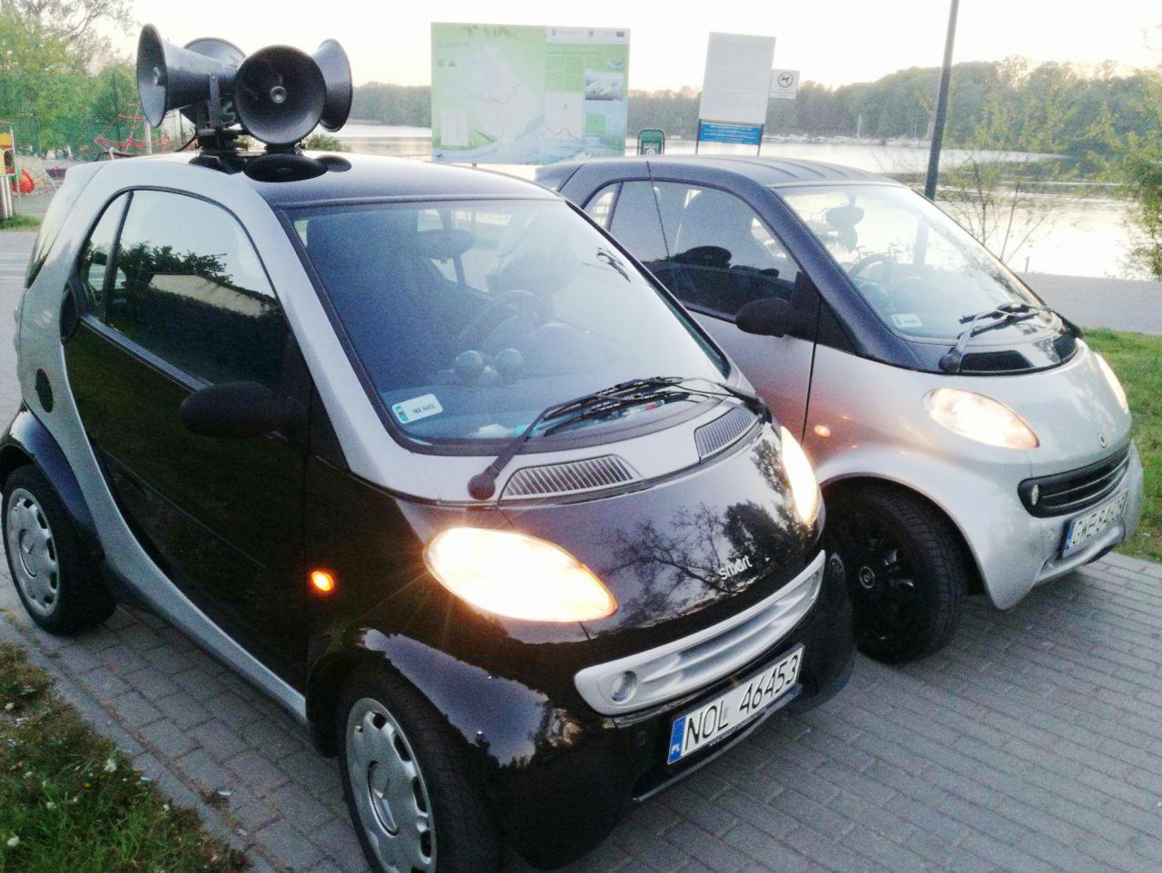 Dwa samochody Smart, jeden czarny z megafonami na dachu, drugi srebrny, zaparkowane obok siebie na tle parku i jeziora.