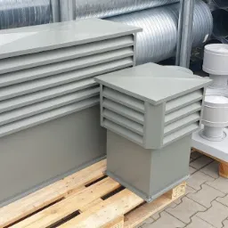 Szare elementy wentylacyjne z poziomymi lamelami, ustawione na paletach na zewnątrz, widoczne rury wentylacyjne w tle.