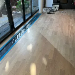 Jasny parkiet przed cyklinowaniem zabezpieczony folią przy oknie tarasowym, w tle odkurzacz przemysłowy i kominek.