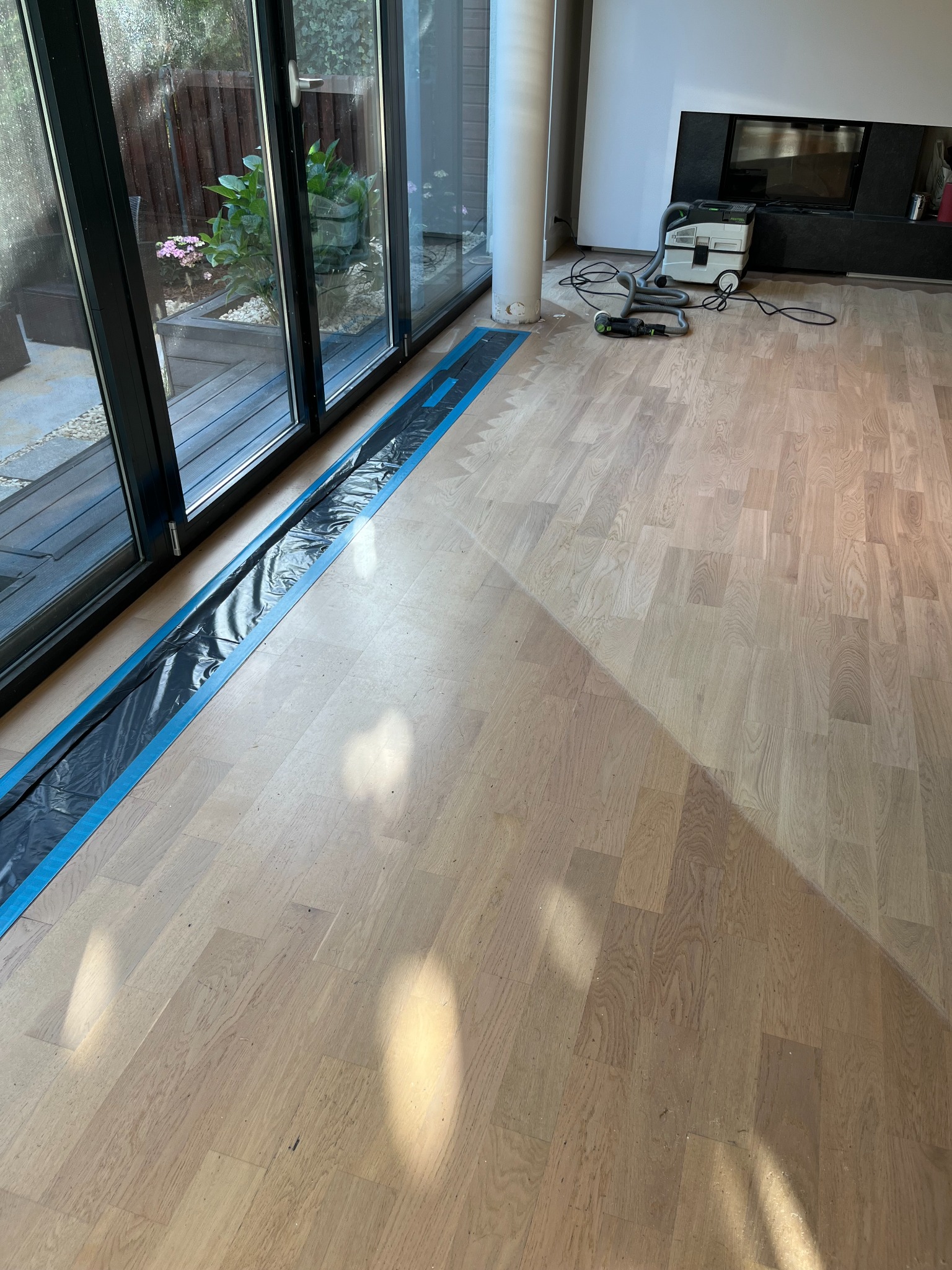 Jasny parkiet przed cyklinowaniem zabezpieczony folią przy oknie tarasowym, w tle odkurzacz przemysłowy i kominek.