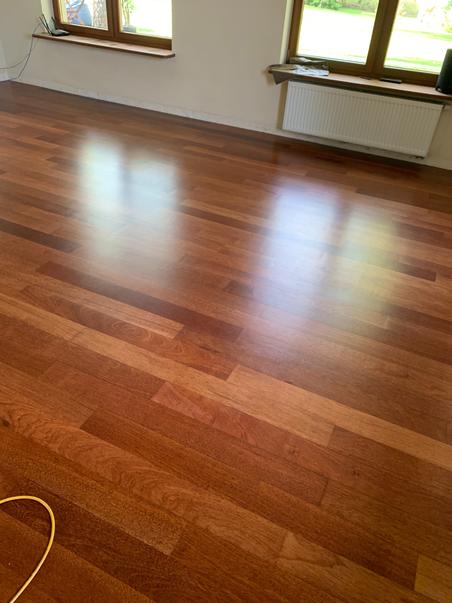 Błyszcząca, świeżo położona podłoga z egzotycznego drewna w pustym pokoju z dwoma oknami, widoczne refleksy świetlne na powierzchni.