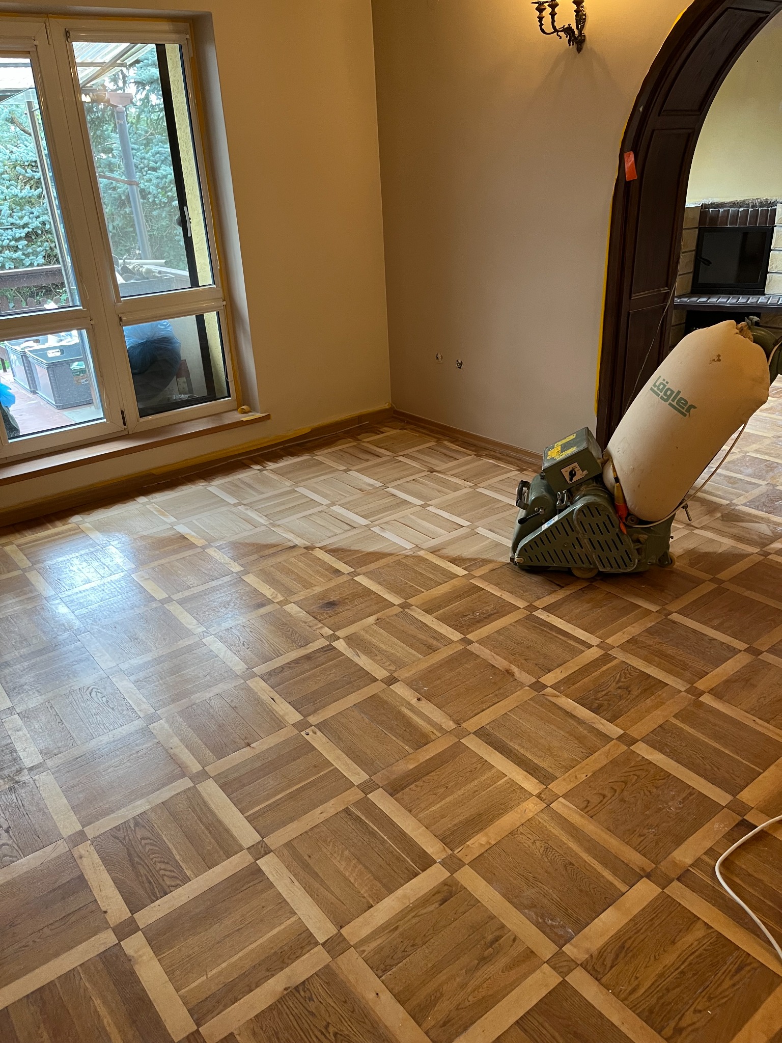 Parkiet w kwadraty częściowo odnowiony, z widoczną maszyną do cyklinowania marki Lägler i taśmą malarską przy oknie.