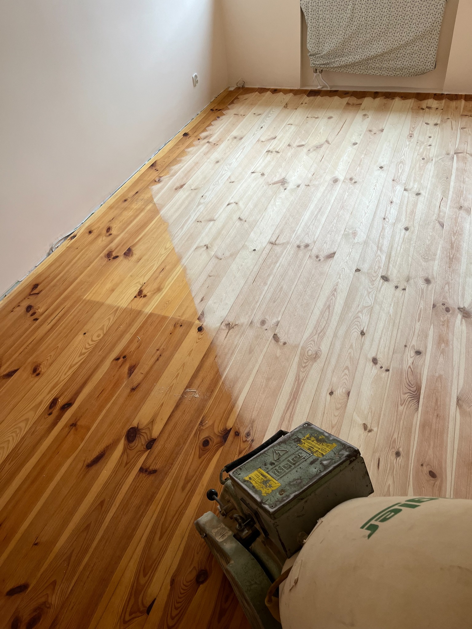 Cyklinowanie drewnianej podłogi z wyraźną różnicą w kolorze między obszarem obrobionym a nieobrobionym, widoczna maszyna do cyklinowania z workiem na pył.