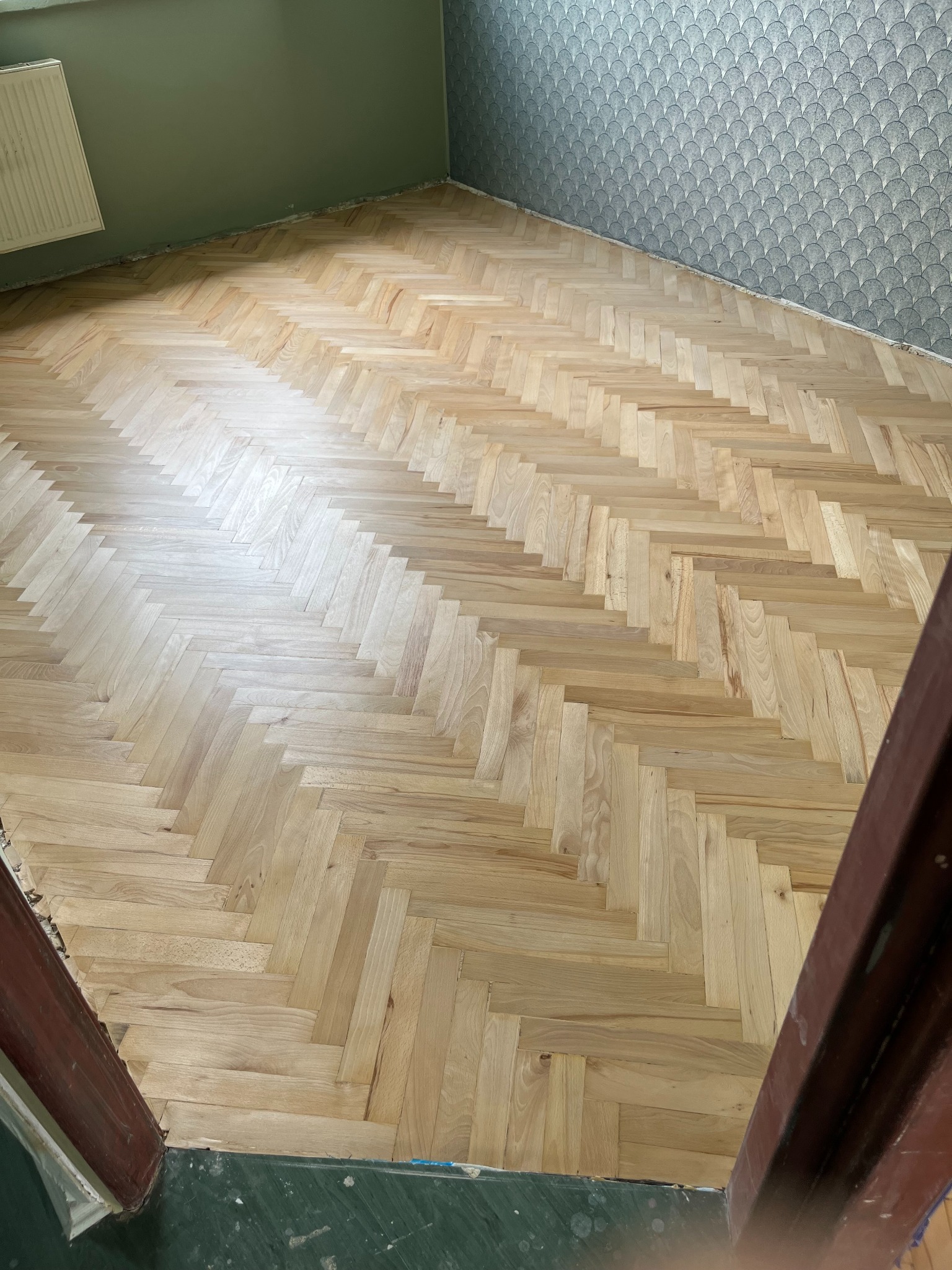 Ułożona jodełka z jasnego drewna parkietowego na podłodze, widoczne niedoskonałości przy ścianach i futrynie, ściana pomalowana na zielono i pokryta tapetą w geometryczny wzór.