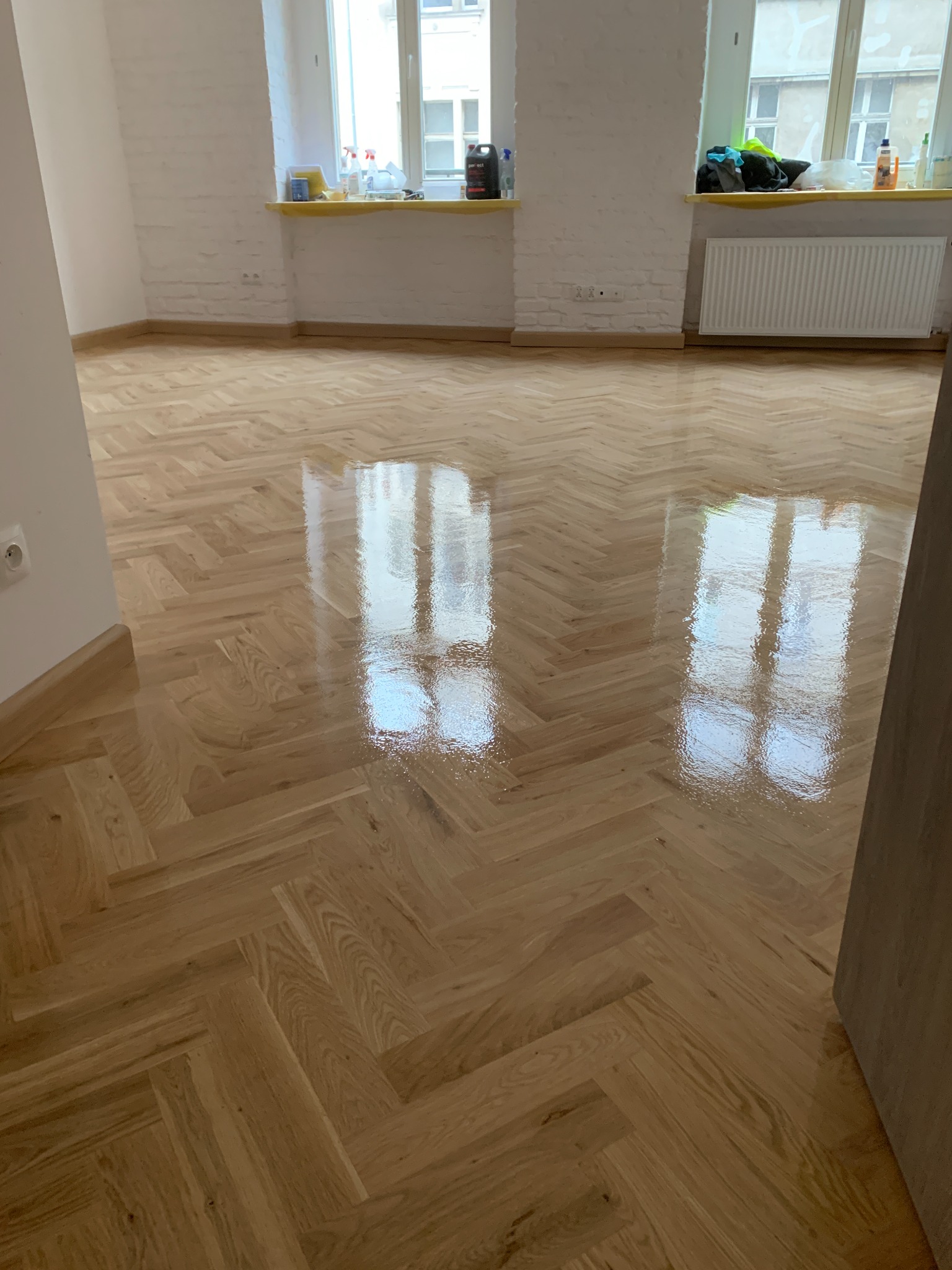 Błyszcząca, nowo położona podłoga z parkietu ułożonego w jodełkę, odbijająca światło z okien, widoczna w pustym, odnowionym pokoju z białymi ścianami.