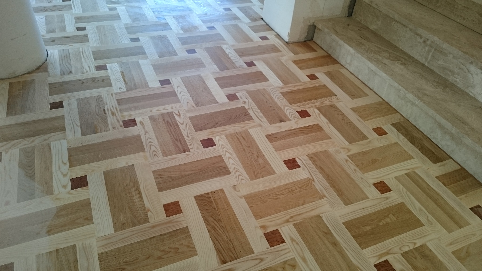 Jasna, drewniana podłoga z parkietu ułożonego w geometryczny wzór z prostokątów i kwadratów, widoczne fragmenty kolumny i marmurowe stopnie schodów.