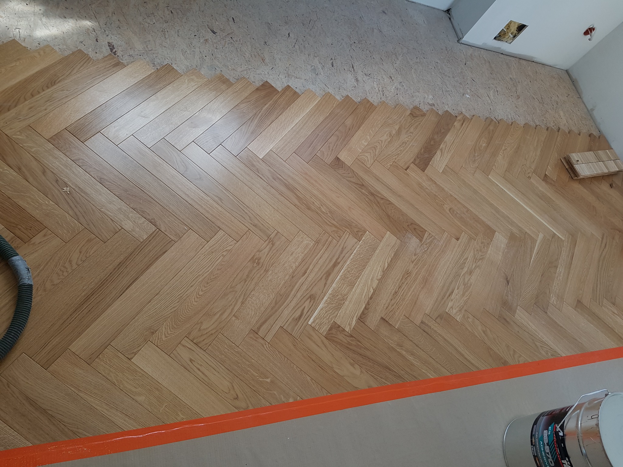 Ułożony parkiet w jodełkę na podłożu, widoczny fragment niezakończonej pracy z nieprzygotowanym podłożem i narzędzia pracy w rogu.