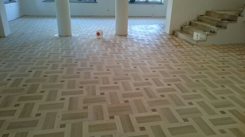 Wzór parkietu ułożonego w jodełkę francuską z ciemniejszymi akcentami, widoczne kolumny w pomieszczeniu i fragment schodów w tle.