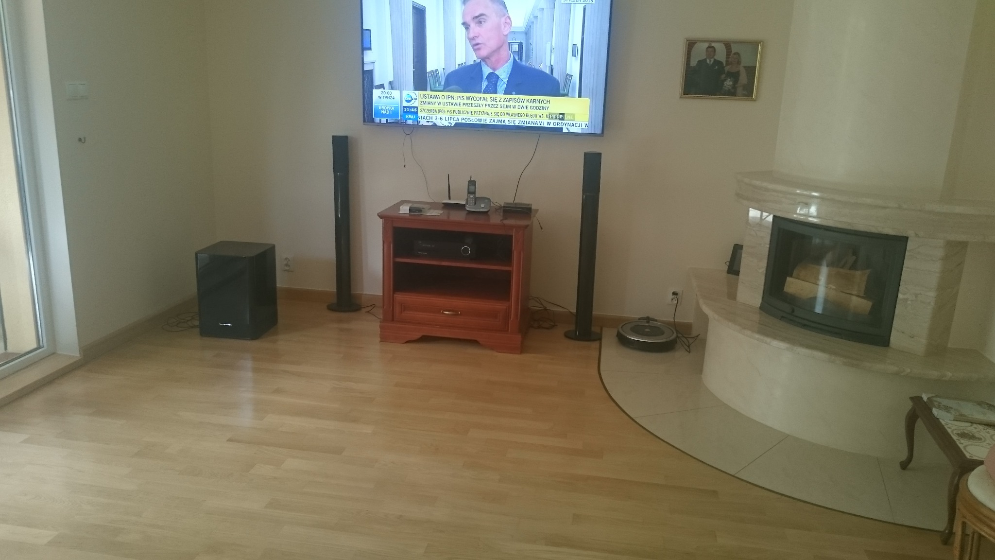 Jasna, lakierowana podłoga z drewna w salonie z kominkiem obłożonym marmurem, widoczne głośniki, szafka RTV, telewizor i robot sprzątający.
