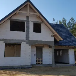 Surowy stan domu murowanego z białych bloczków, widoczna konstrukcja dachu z drewnianych belek i czarna dachówka, w tle las iglasty.