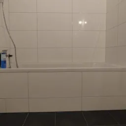 Wykończona łazienka w Mszanie Dolnej z białą wanną obudowaną płytkami, grzejnikiem drabinkowym po lewej i czarną podłogą z płytek. Na wannie stoją kosmetyki.