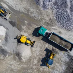 Zdjęcie z lotu ptaka przedstawia teren budowy z ciężarówką wywrotką, dwiema koparkami i ładowarką na tle hałd żwiru i piasku.