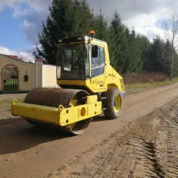 Żółty walec drogowy Stalzlom BW 177DH-3 ubija gruntową drogę, widoczne ślady opon, w tle drzewa i ogrodzenie posesji.