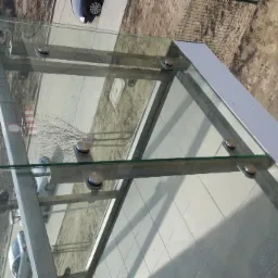 Szklana konstrukcja z mocowaniami punktowymi, widok z góry, z elementami otoczenia w tle, w tym fragmentem parkingu i zieleni.