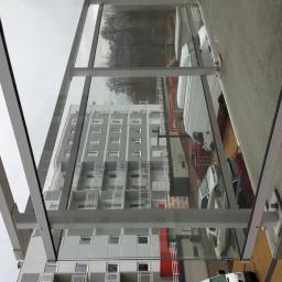 PHU PROFIL-TECH Grzegorz Kuźlik - Szklane zadaszenie nad parkingiem, widok na budynek mieszkalny i zaparkowane samochody w tle, metalowa konstrukcja wspierająca.