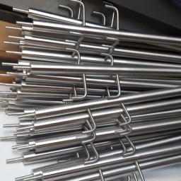 PHU PROFIL-TECH Grzegorz Kuźlik - Stos polerowanych, nierdzewnych rurek z wygiętymi uchwytami na końcach, ułożonych warstwami na białej powierzchni, w tle widoczny fragment żółtej ściany i niebieska szczotka.