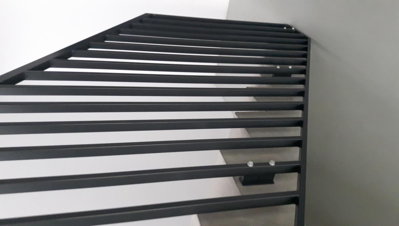 Minimalistyczna, czarna, metalowa konstrukcja schodów z poziomymi szczeblami, mocowana do białej ściany i szarej ściany, widok z dołu.