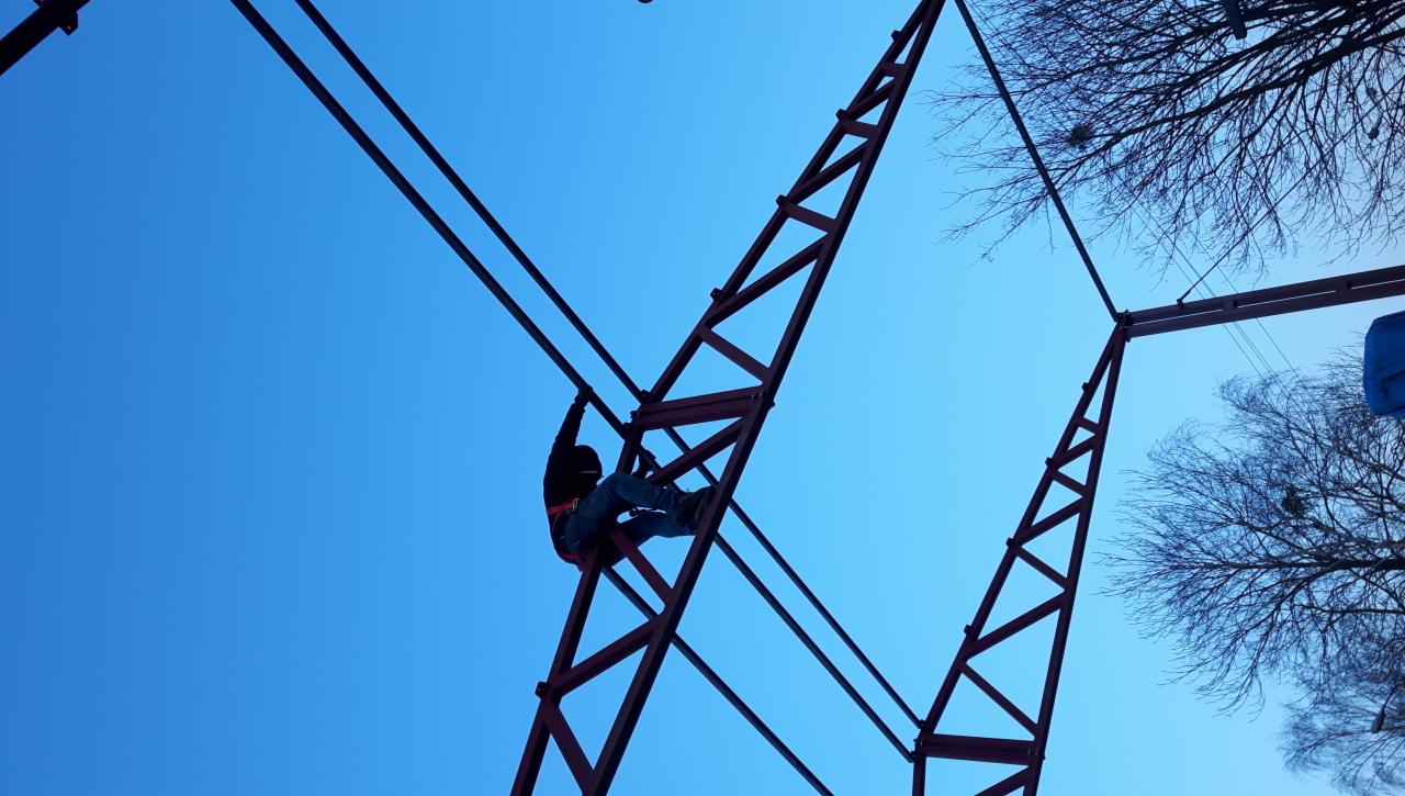 Pracownik w uprzęży asekuracyjnej wspina się po metalowej konstrukcji, prawdopodobnie w trakcie montażu lub naprawy, na tle błękitnego nieba.