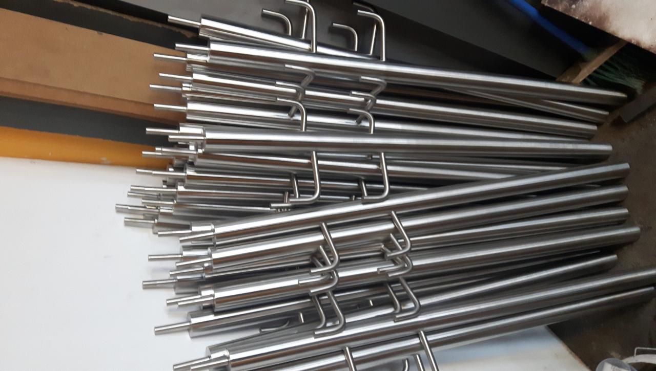 Stos polerowanych, nierdzewnych rurek z wygiętymi uchwytami na końcach, ułożonych warstwami na białej powierzchni, w tle widoczny fragment żółtej ściany i niebieska szczotka.