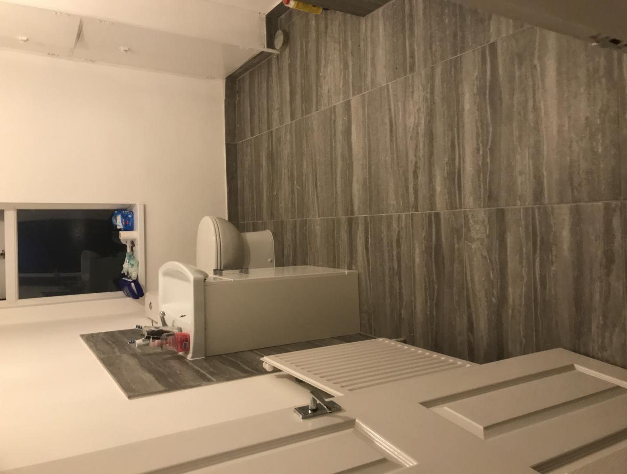 Fragment łazienki w trakcie remontu: widoczna toaleta, umywalka, płytki imitujące drewno na ścianie oraz białe, częściowo zdemontowane drzwi.