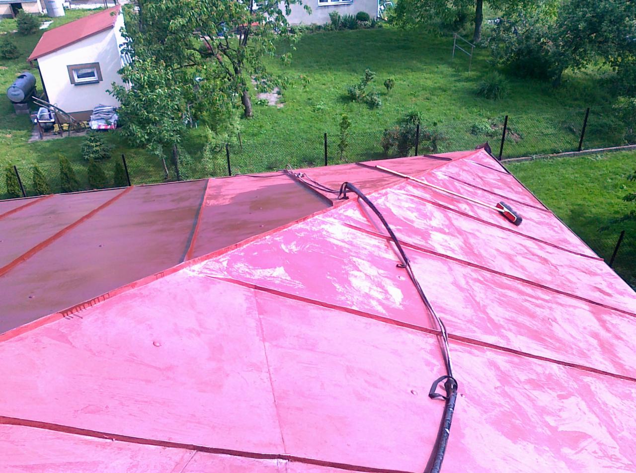 Czerwono-różowy dach z blachy trapezowej po umyciu, z widocznym wężem ciśnieniowym i szczotką do czyszczenia, na tle zielonego trawnika i zabudowań.