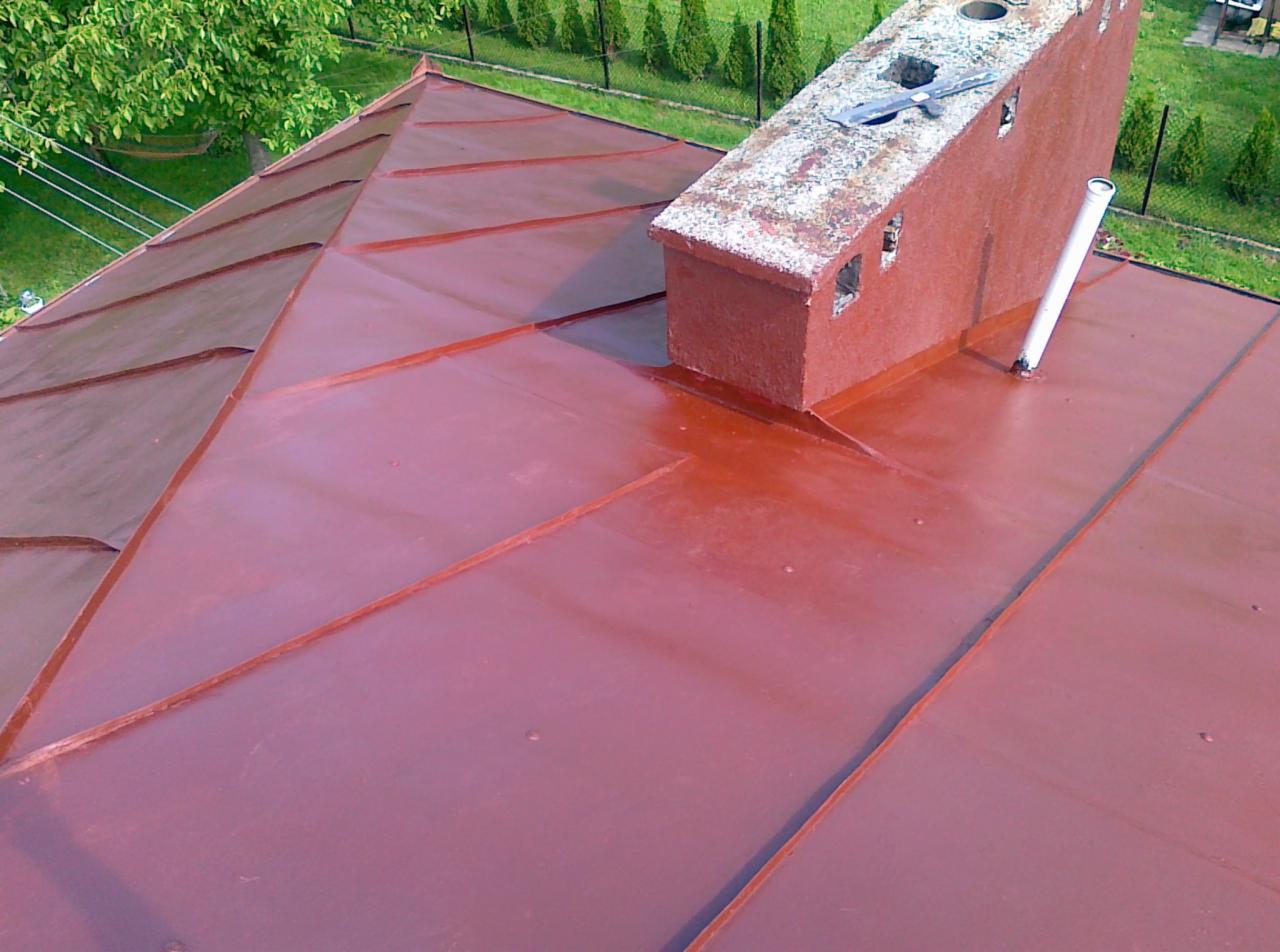 Czerwono-brązowy, metalowy dach z widocznym kominem i rurą wentylacyjną, sfotografowany z góry, z zielonymi drzewami w tle.