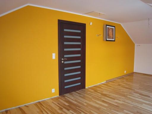 Pokój na poddaszu po odświeżeniu: ściany pomalowane na intensywny żółty kolor, brązowe drzwi z poziomymi przeszkleniami, parkiet na podłodze, obraz na ścianie.
