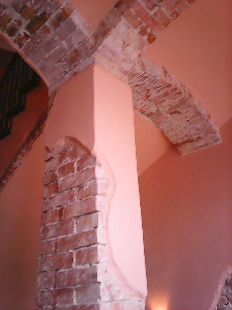 Ceglany łuk i filar w trakcie wykańczania, część ściany pomalowana na różowo, widoczne ślady prac przygotowawczych.