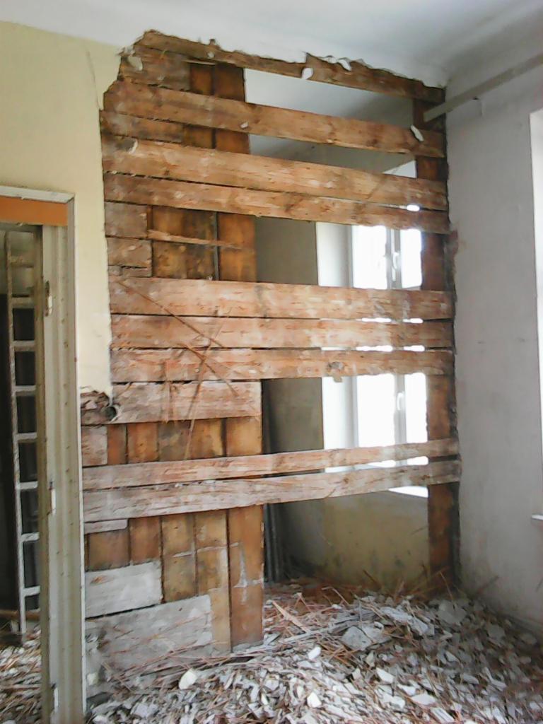 Demontaż ściany z drewnianą konstrukcją nośną, gruz i resztki tynku na podłodze, widok na okno w tle.