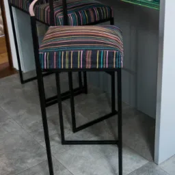 Dwa nowoczesne hokery z czarną metalową ramą i tapicerowanymi siedziskami w kolorowe paski, ustawione przy blacie w jasnym wnętrzu.