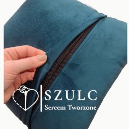Anna Szulc - Szmaragdowa poszewka na poduszkę z wszytym zamkiem błyskawicznym, częściowo rozpiętym przez dłoń z beżowym lakierem do paznokci, widoczne logo firmy.