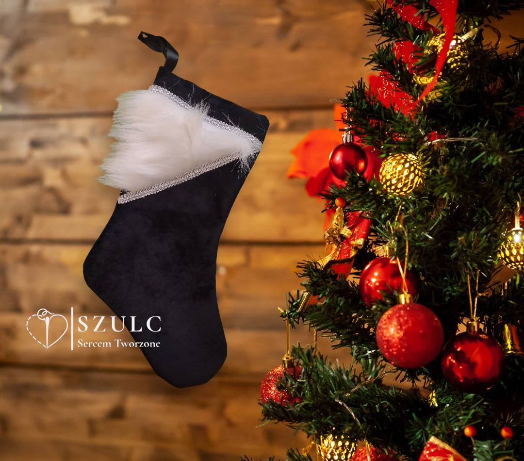 Czarna, aksamitna skarpeta świąteczna z białym futerkiem i srebrną lamówką, wisząca obok choinki z czerwonymi i złotymi bombkami, logo firmy widoczne w lewym dolnym rogu.