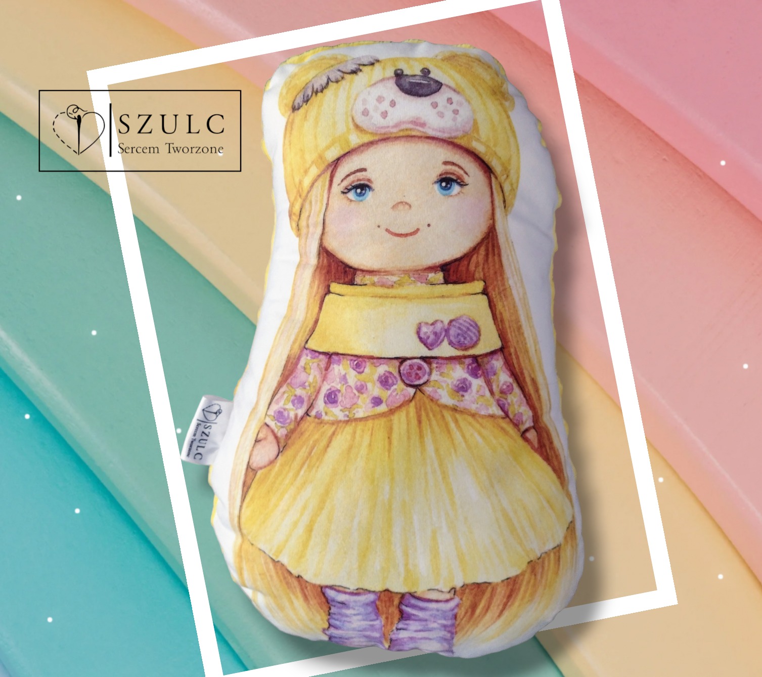 Dekoracyjna poduszka w kształcie lalki z blond włosami, w żółtej sukience i czapce z motywem misia, prezentowana na tle w pastelowych kolorach.