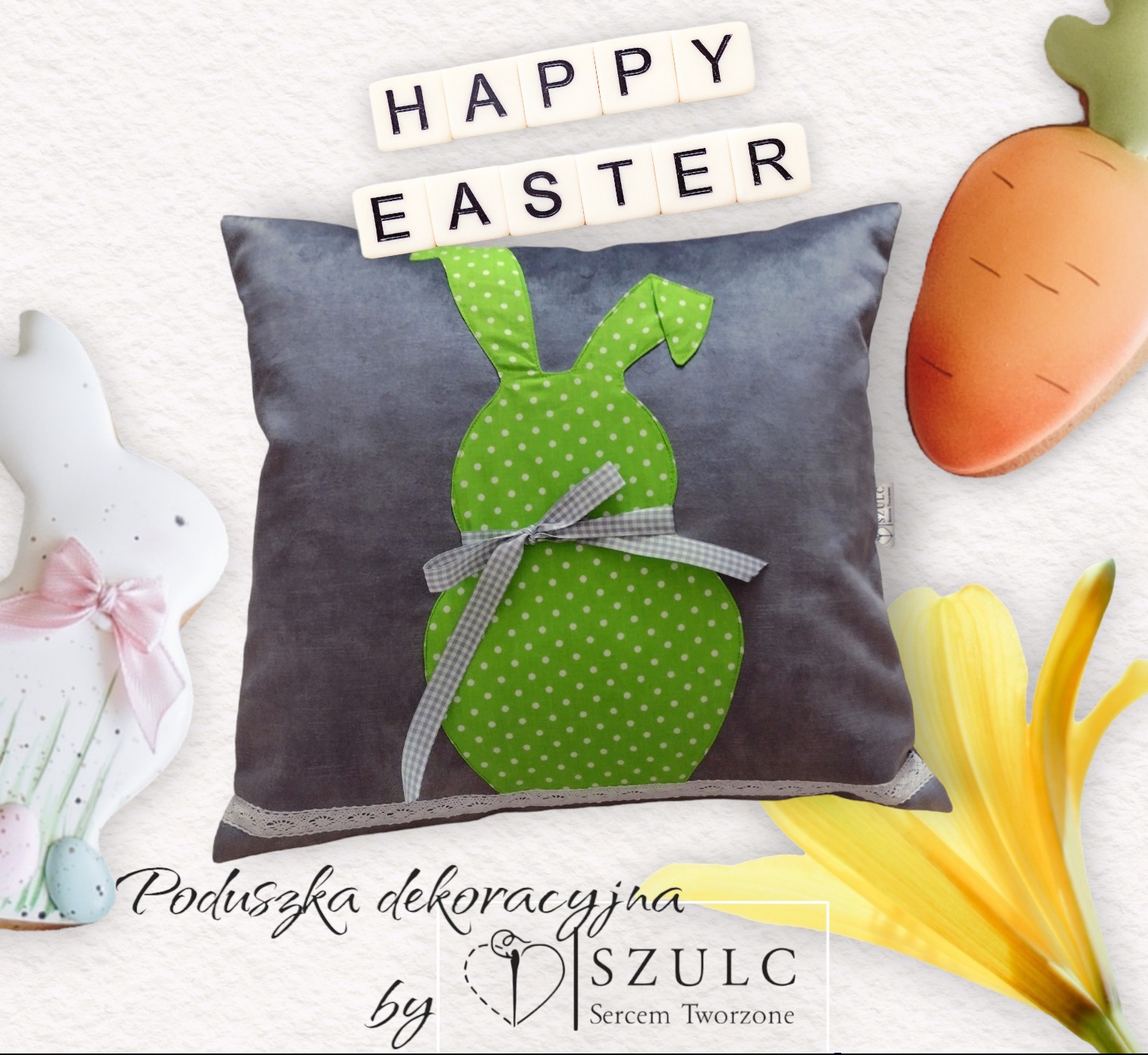Dekoracyjna szara poduszka z aplikacją zielonego zajączka w białe kropki, przewiązana szarą wstążką w kratkę, otoczona wielkanocnymi dekoracjami: ciasteczkowy zajączek z różową kokardką, marchewka...