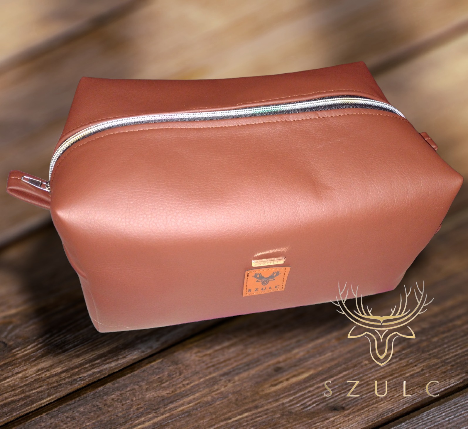 Brązowa, prostokątna kosmetyczka z zamkiem błyskawicznym, wykonana z imitacji skóry, z małą metką z logo firmy na tle drewnianego blatu.