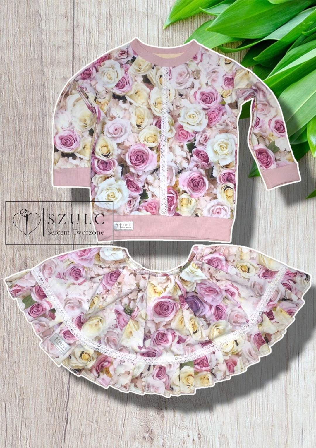 Dziecięcy komplet: bluza z długim rękawem i spódniczka z falbanką, obie uszyte z materiału w róże, wykończone białą koronką, na drewnianym tle z zielonymi liśćmi. Widoczne logo producenta.