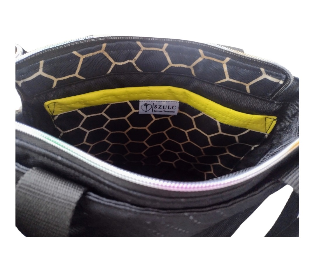 Wnętrze czarnej torby z wzorem plastra miodu, wykończone żółtą lamówką, widoczne logo 'SZULC Sercem Tworzone' na kieszeni.