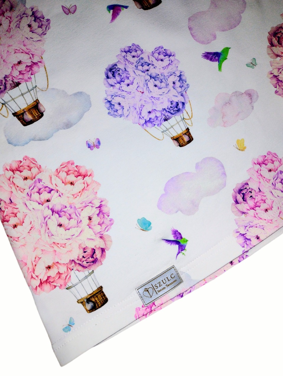 Fragment białej tkaniny z nadrukiem akwarelowych balonów z kwiatów, chmur, motyli i kolibrów, widoczny narożnik materiału i metka z logo.