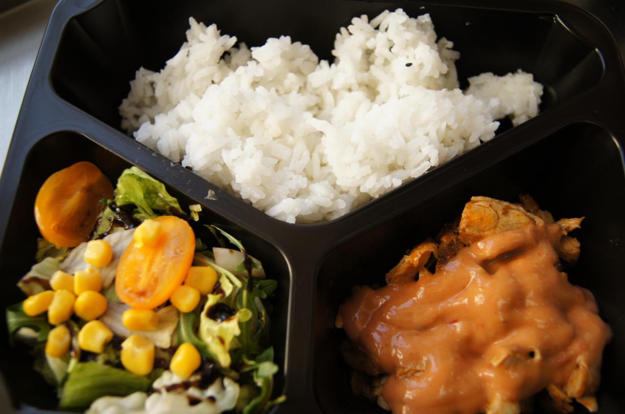 Czarny pojemnik na wynos podzielony na trzy części: ryż, sałatka z kukurydzą i pomidorami koktajlowymi polana sosem balsamicznym, oraz mięso w sosie pomidorowym.